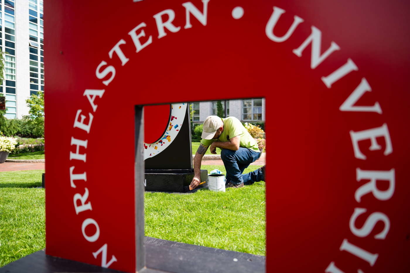 Framed by a sign, a person paints the corner of another sign outside on grass.