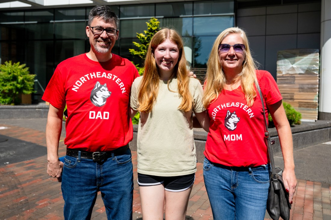 A Northeastern student stands smiling between her parents, who proudly wear red “Northeastern Dad” and “Northeastern Mom” shirts, outside the East Village dormitory.