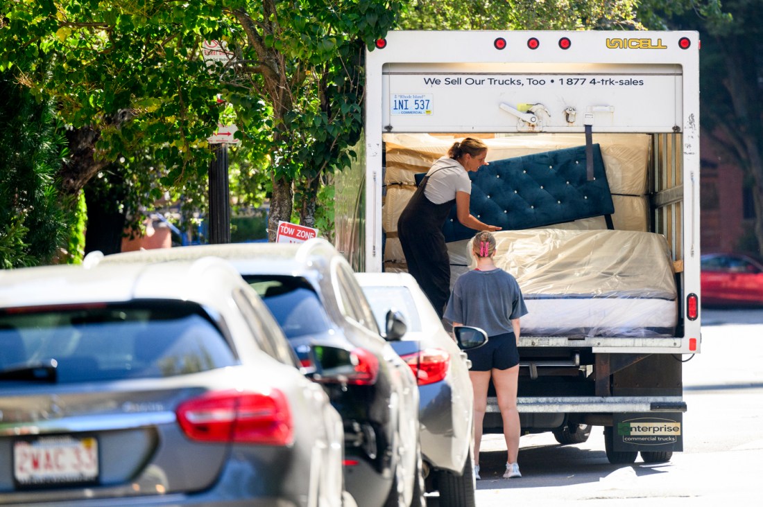 A person in dark colored overalls and a white t-shirt taking a blue tufted headboard out of the back of a white moving truck.