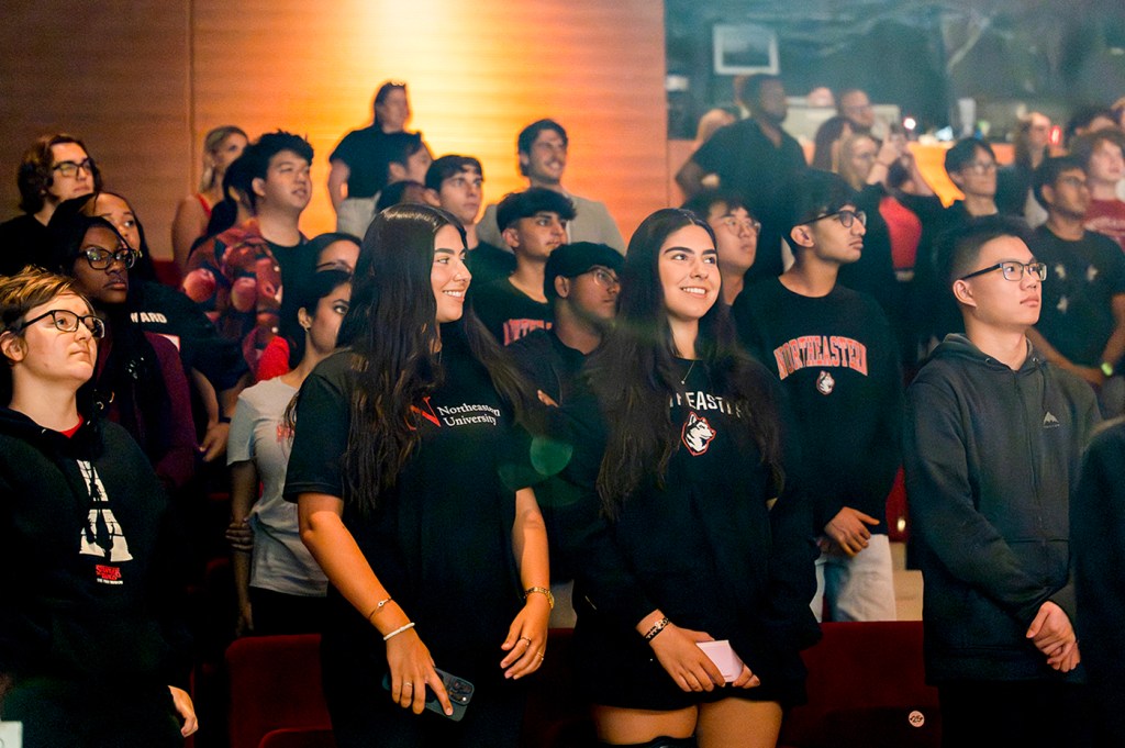 A diverse group of students sits in rows at the NYC Convocation ceremony, attentively watching the stage. Many wear university sweatshirts and casual clothing.