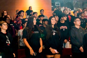 A diverse group of students sits in rows at the NYC Convocation ceremony, attentively watching the stage. Many wear university sweatshirts and casual clothing.