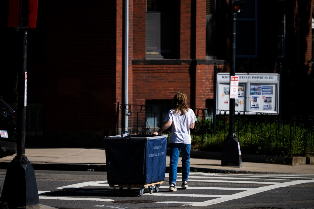 A person in a white t-shirt and jeans walks away from the camera while pulling a wheeled bin toward a red brick building with large windows.
