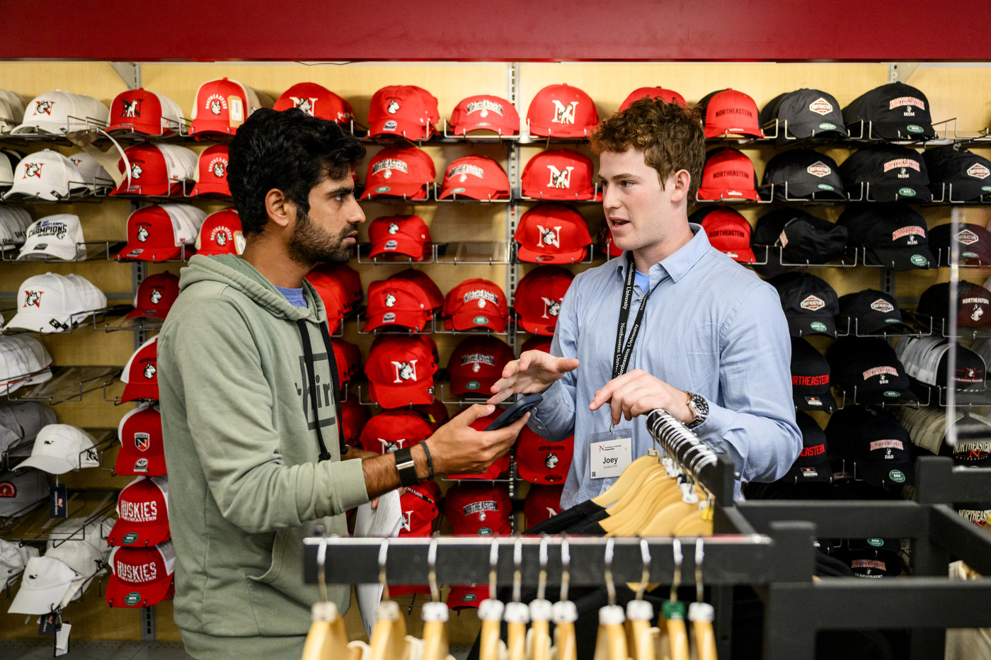 Joe Friedman depicted in a blue shirt helping another student by a shelf of hats.