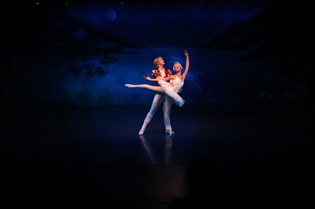 A ballet pas de deux performed on a dark stage with deep blue atmospheric lighting. A male dancer in a burgundy velvet tunic supports a female dancer in a white classical tutu as she executes an arabesque en pointe, with one leg extended behind her and one arm gracefully raised upward.