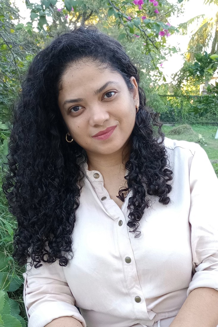 Author Abigail Persaud Cheddie smiles softly outdoors, wearing a cream blouse with gold hoop earrings, her long curly hair over one shoulder, with green trees and pink flowers behind her.