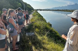 A group of students stand on a grassy path looking at a river. An instructor stands in the foreground.