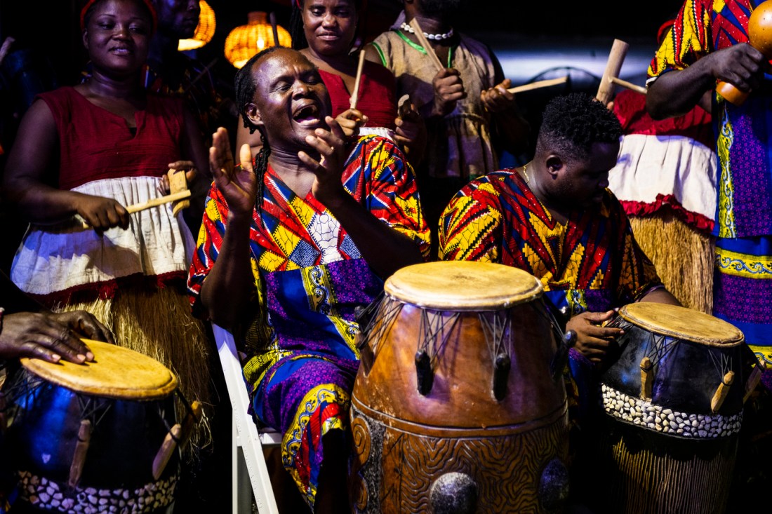 A group of Black drummers dressed in traditional garb clap and drum together.