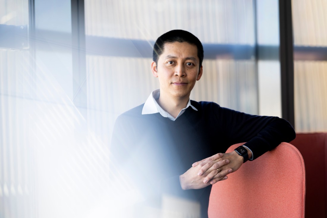 Jing-Ke Weng, Northeastern professor, sits in a modern campus building with natural light streaming through the windows, hands folded, and wearing a dark sweater and smart watch.