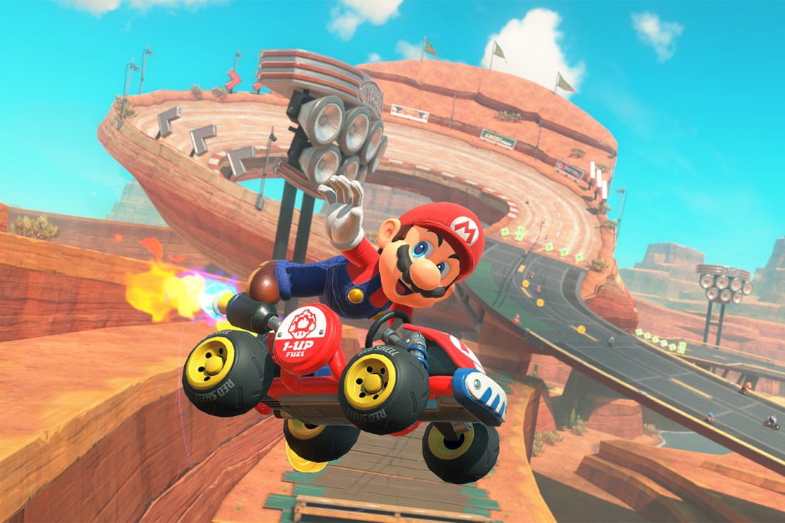 A screen capture of the Mario character in a race car floating in the area over a race track that extends into the background.