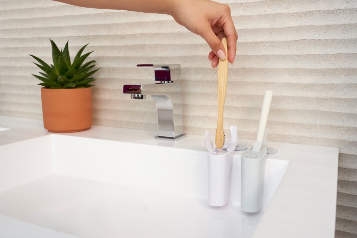 A hand lifts a bamboo toothbrush from a stand next to a bathroom sink, with a potted succulent on the counter nearby.