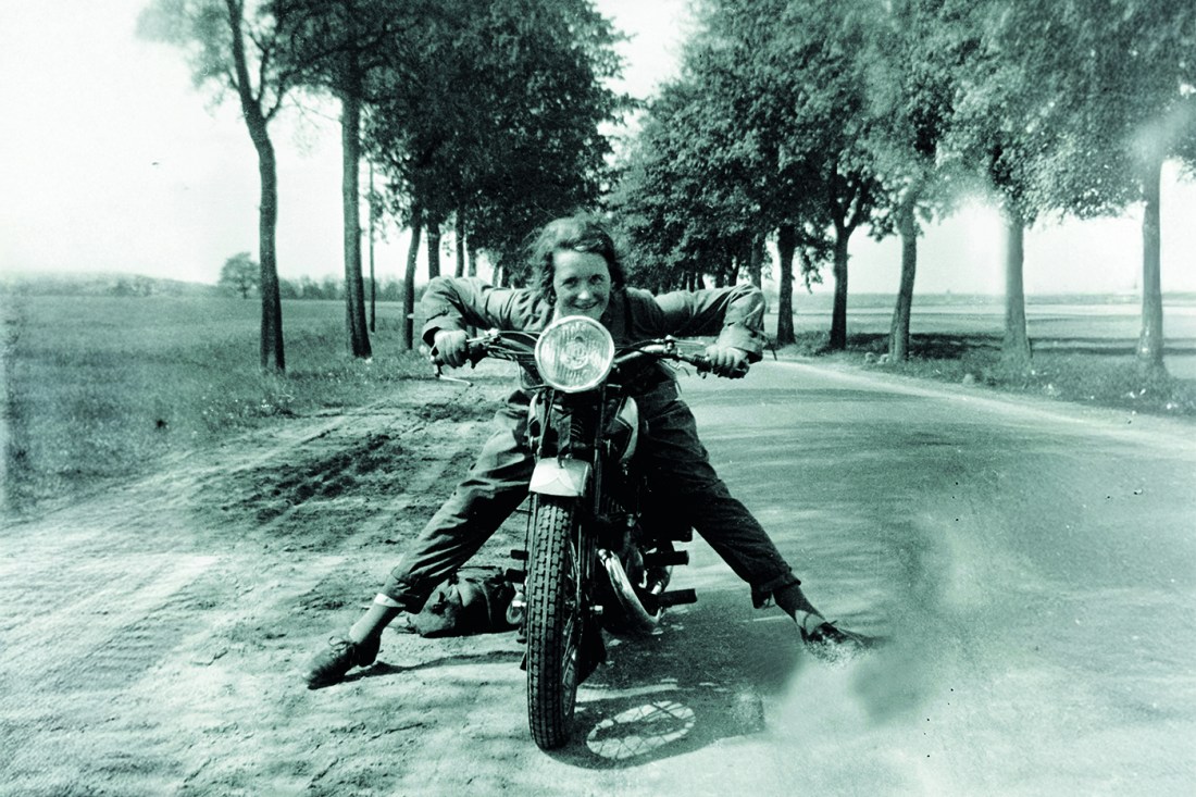 A smiling woman holding the handlebars of her motorcycle; feet on either side of the wheels as she sits on it. She's smiling with excitement and perhaps mischievousness into the camera. Behind her is a dirt tree-lined road.