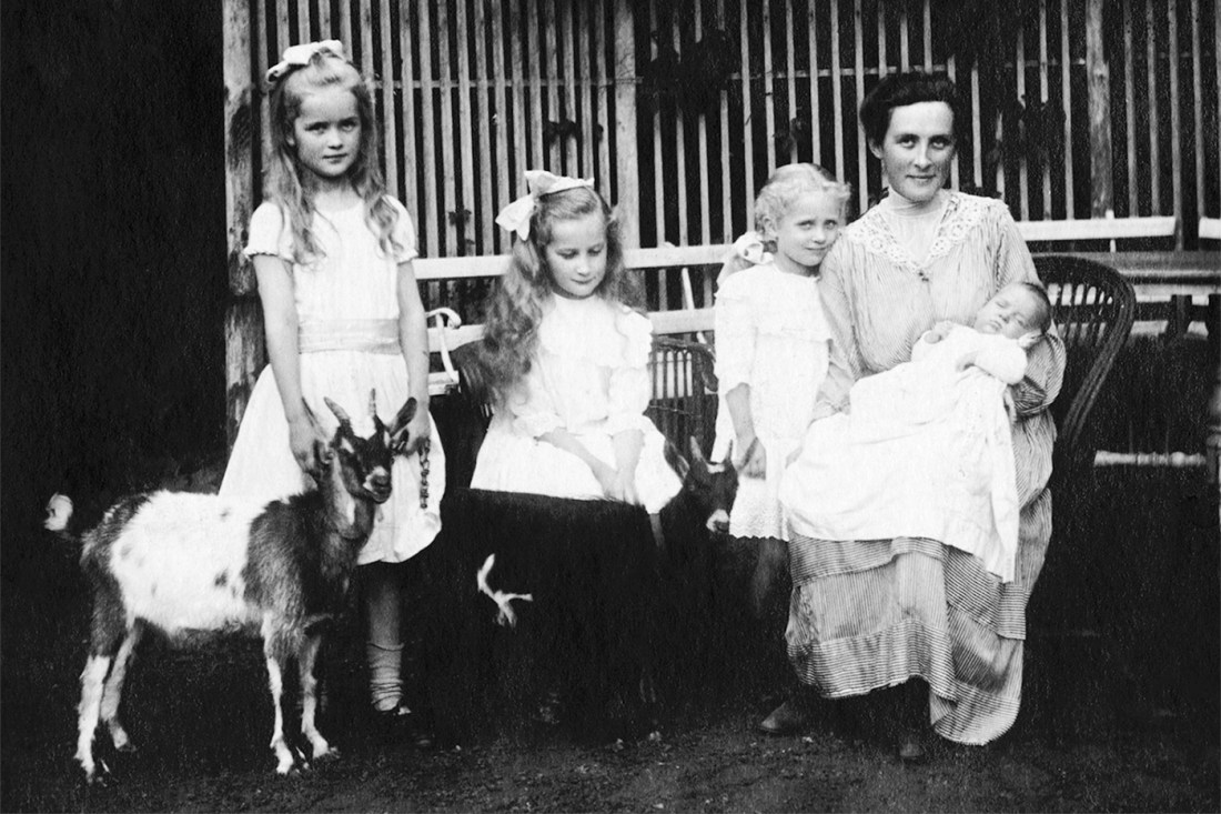 A black-and-white photo of a parent with a baby in arms, and 3 small children nearby. The oldest child holds a goat.