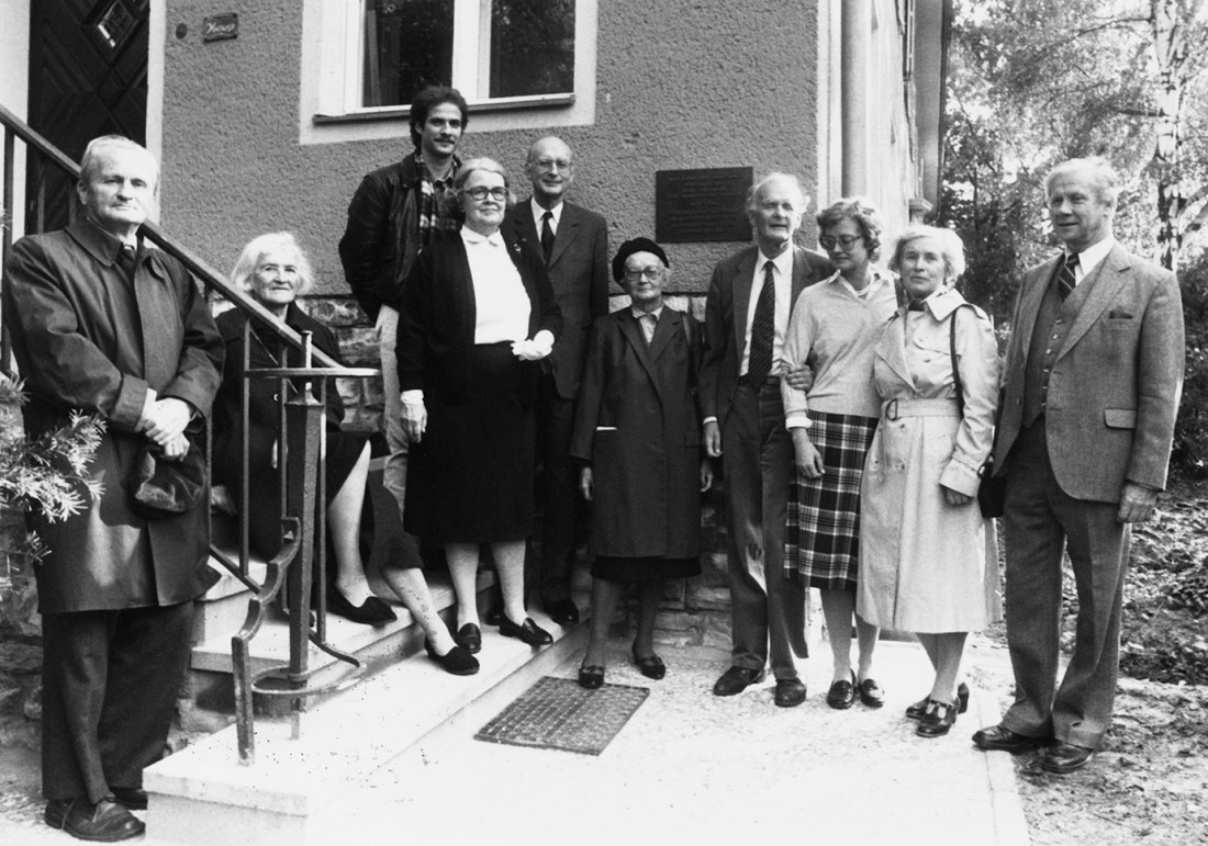 A group photo of ten adults of varying ages standing outside. Everyone is smartly dressed. Some stand or sit on the steps of a family home.