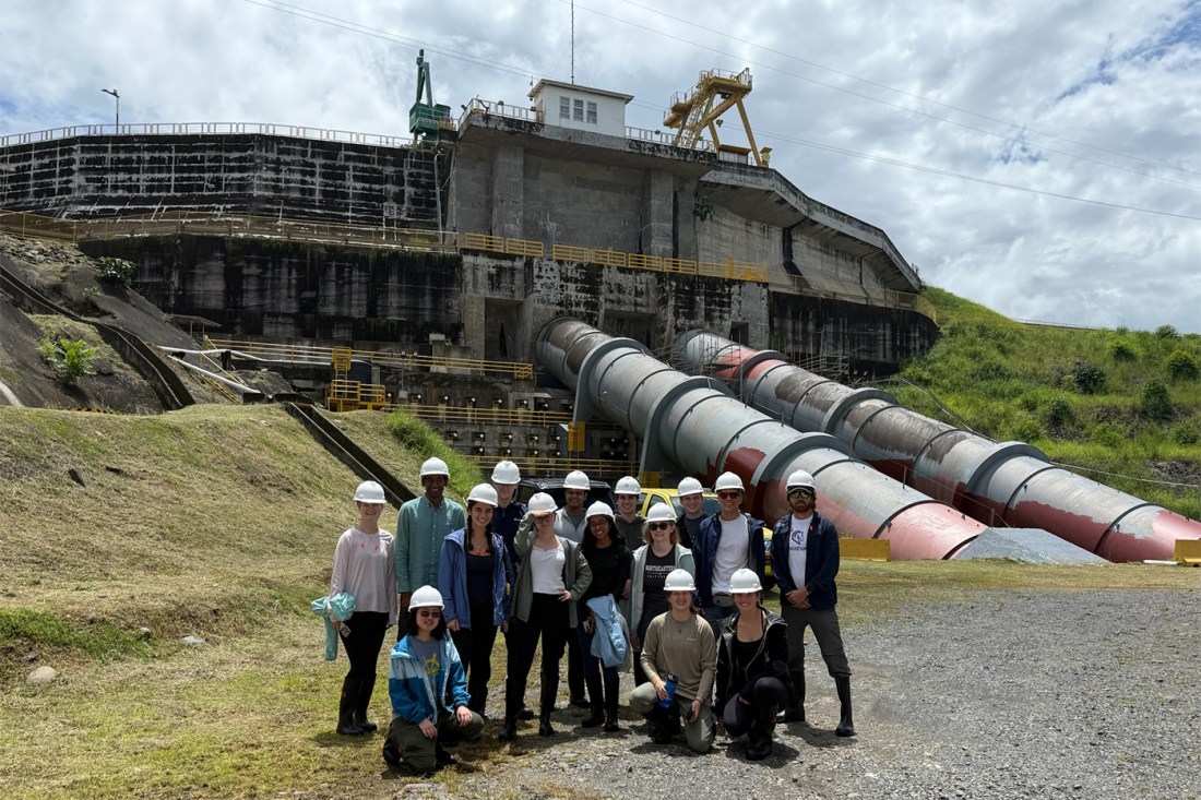 A group of people wearing white hard hats and safety gear pose for a group photo in front of a large industrial facility at the Panama Canal.