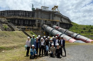 A group of people wearing white hard hats and safety gear pose for a group photo in front of a large industrial facility at the Panama Canal.