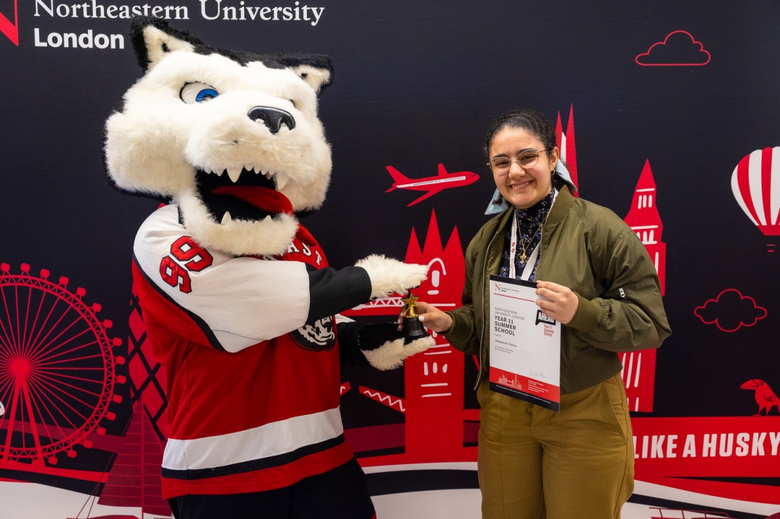 Paws gives a small trophy to a smiling student who holds a paper saying Year 11 Summer School. Behind them is a backdrop of graphical elements representing London.