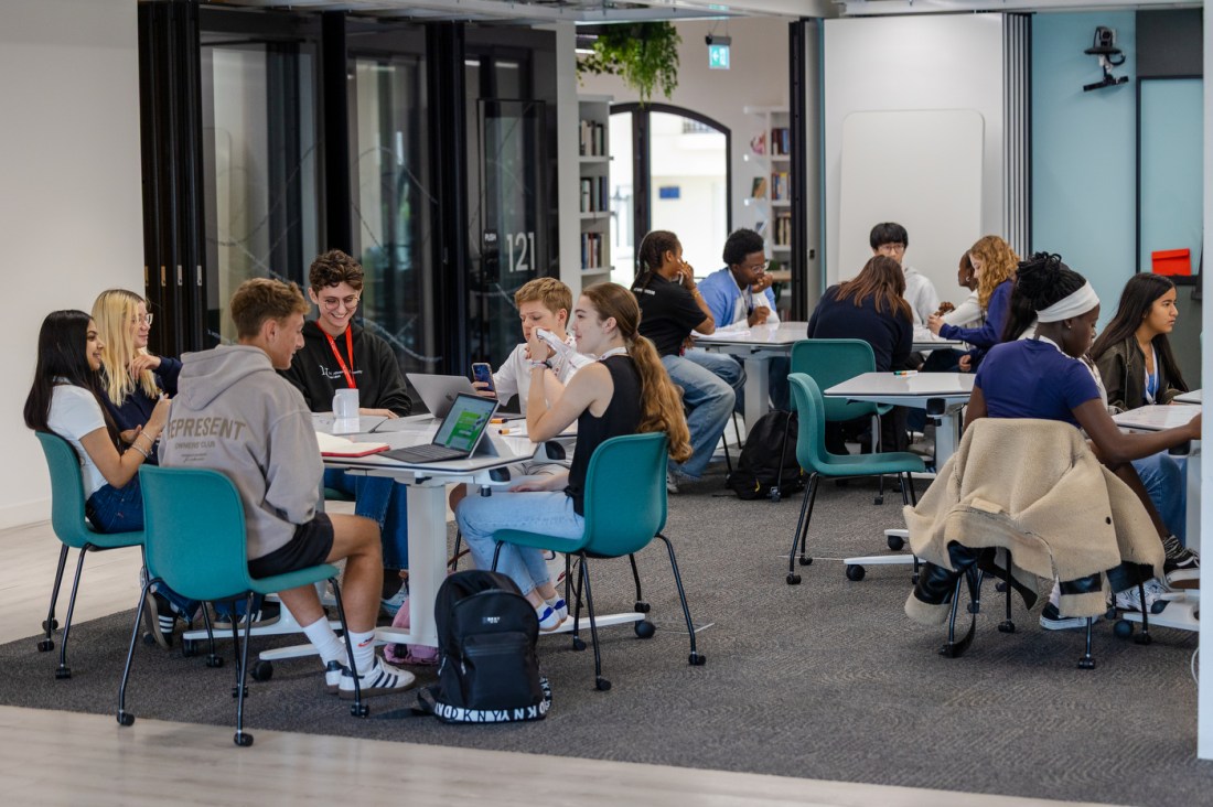 Groups of students sit at large tables chatting and working together.