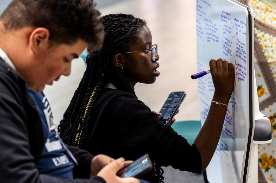 A student writing on a small whiteboard with a purple marker while another looks something up on their phone.