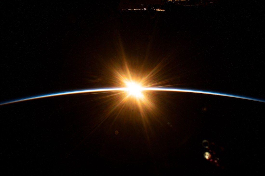 A photo of an eclipse. The sun rises over the Earth’s horizon, as seen from space, casting a golden glow against the darkness of space. A thin blue curve marks the planet’s atmosphere.