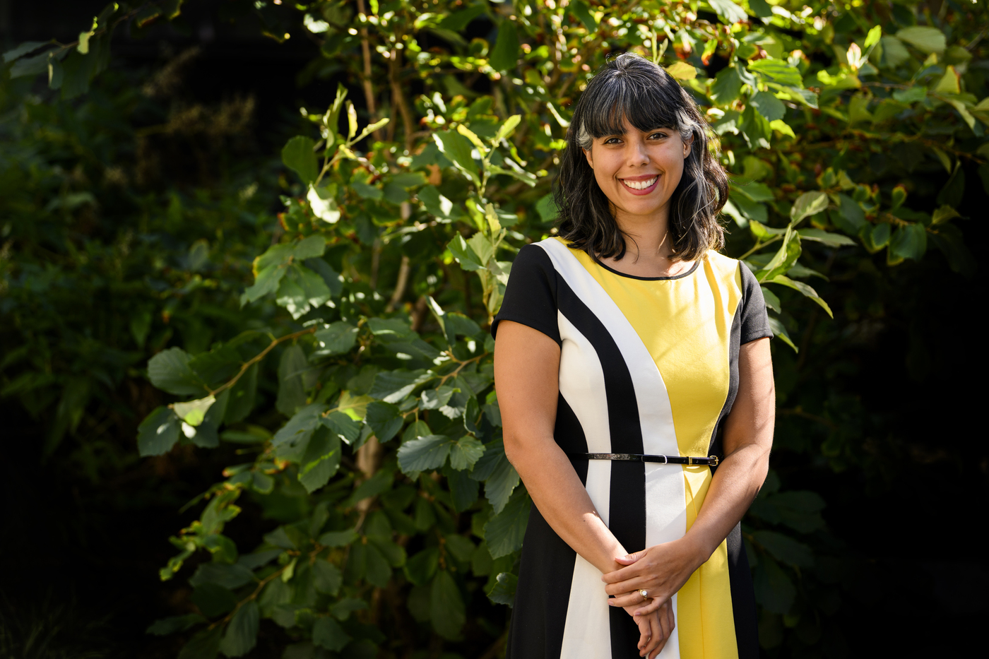Portrait of Stephanie Noble, smiling in front of greenery, wearing a yellow, black, and white dress.