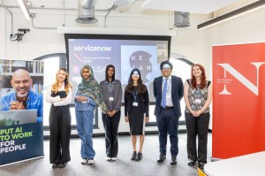 Six graduates of Northeastern University’s fast-track digital course pose side by side in a classroom, smiling in front of a projector screen and banners for ServiceNow and Northeastern. The cohort includes men and women in professional attire, representing a diverse group.