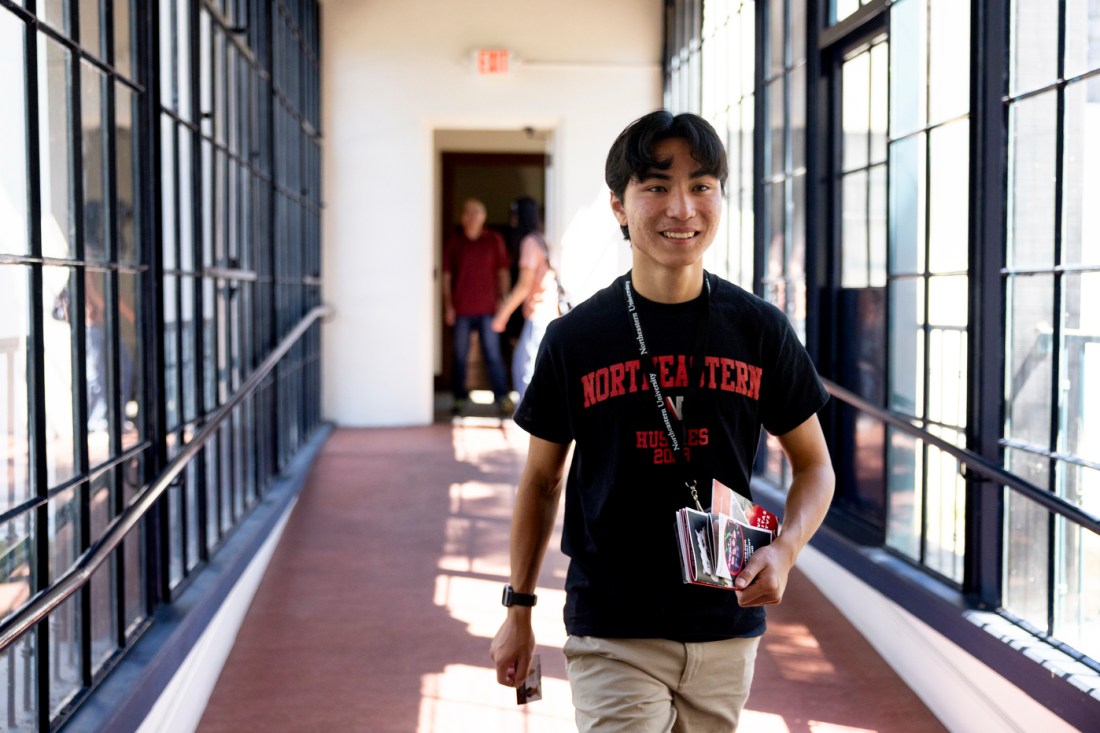 A student wearing a Northeastern shirt and a lanyard walks down a hallway carrying several pamphlets in their hand.