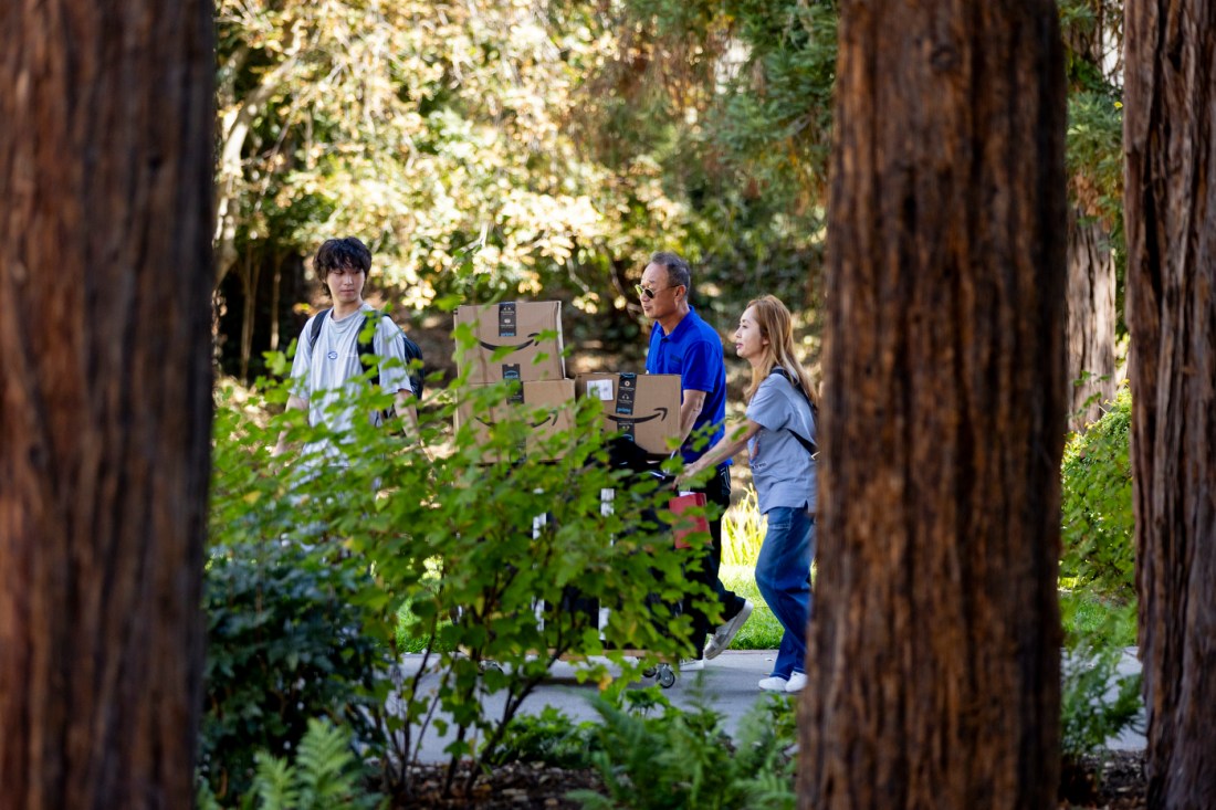 A family pushes a cart stacked with Amazon boxes along a walkway.