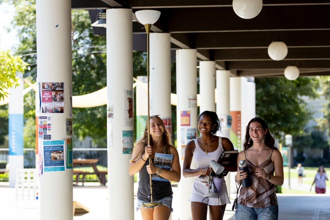 Three students walk under a walkway with hanging globe lights and white pillars. One of the students is carrying a tall lamp.