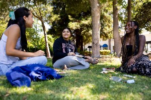 Three female students sit cross-legged on grass under trees at Northeastern University's Oakland campus, having a casual outdoor conversation during a sunny day.