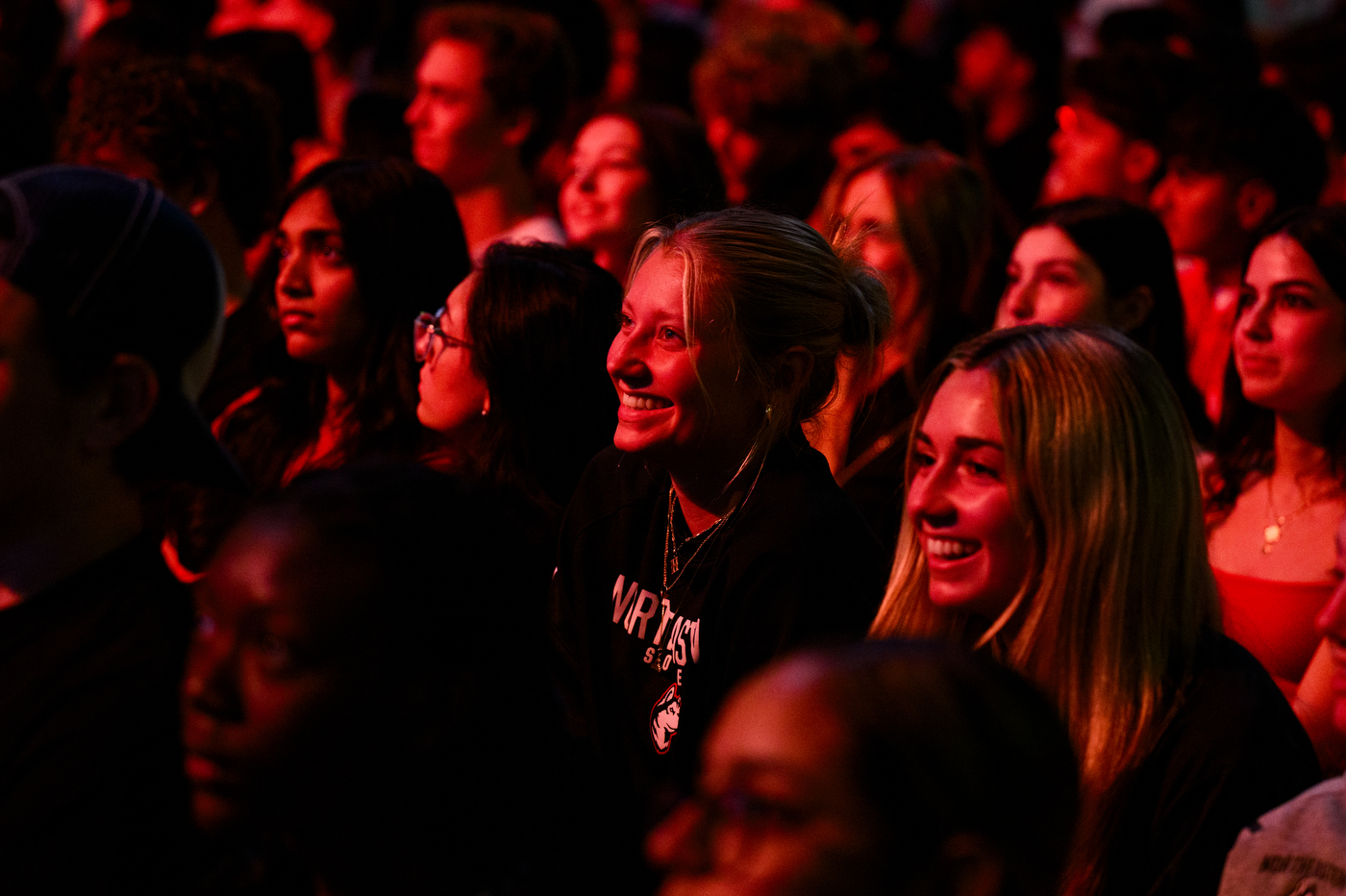 Students in the audience smile and watch the event closely, illuminated by a red light.