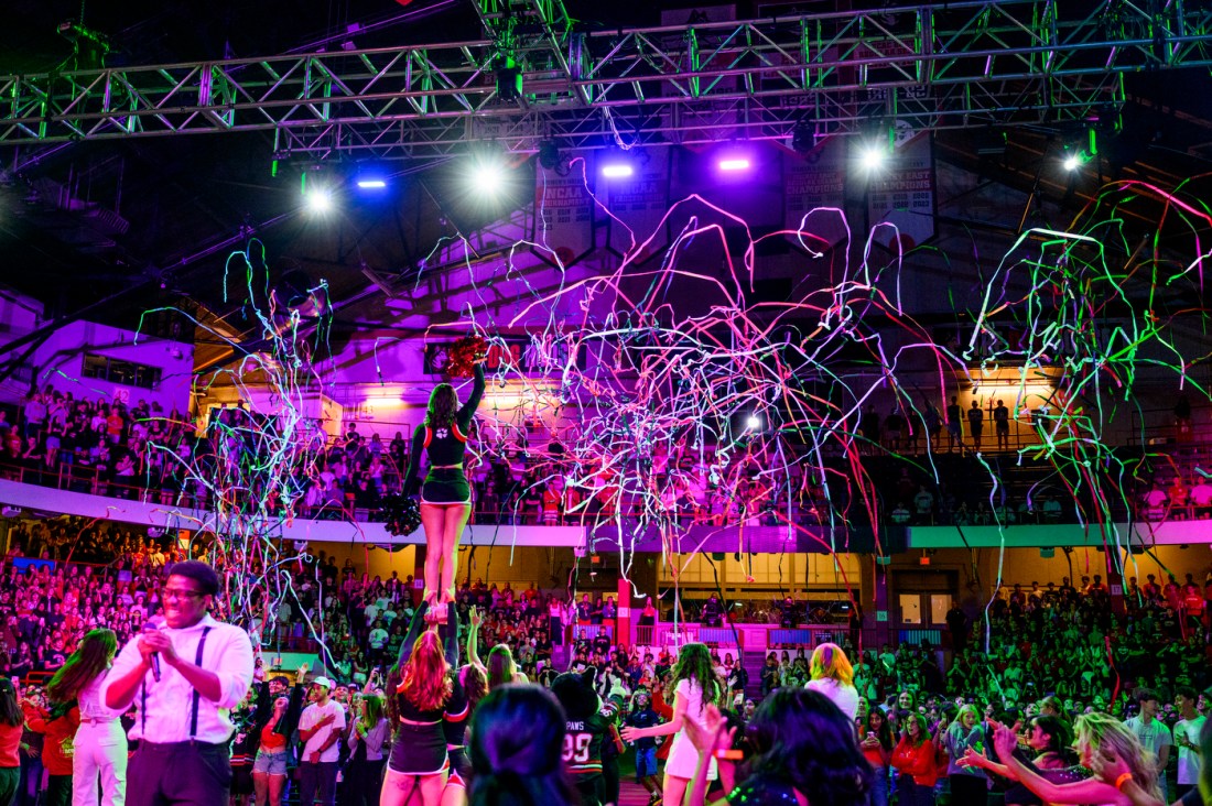 Confetti streams through the air as cheerleaders perform in front of a packed arena with colorful lights overhead.