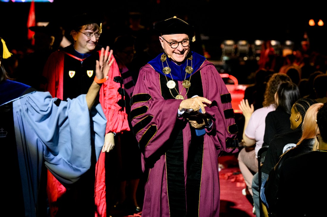 A university official in academic regalia smiles and clasps hands while walking through a crowd of students at convocation.