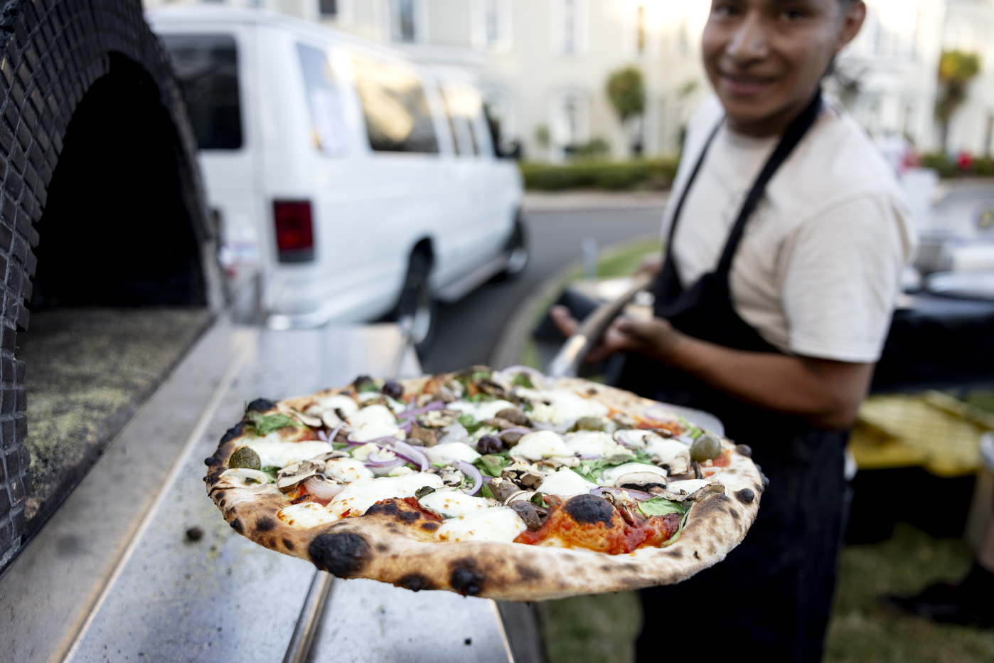 A person wearing a white t-shirt under a black apron holds a paddle with a pizza on it up to the camera.