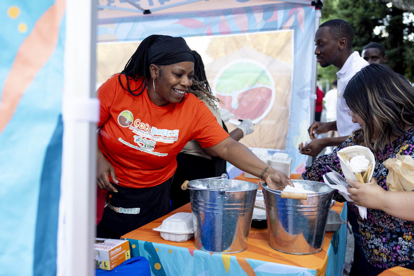 A person wearing an orange t-shirt helping serve another person food from a large silver pot.