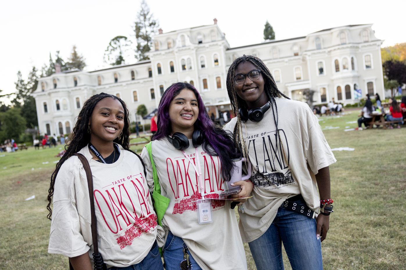 Three smiling Northeastern University students stand on a grassy lawn in front of a large cream-colored Victorian-style building at the Oakland campus. All three are wearing oversized “Taste of Oakland” T-shirts with bold red or black lettering, headphones around their necks, and ID badges. They appear relaxed and cheerful during Fall Orientation Week, with other students and picnic tables scattered in the background.
