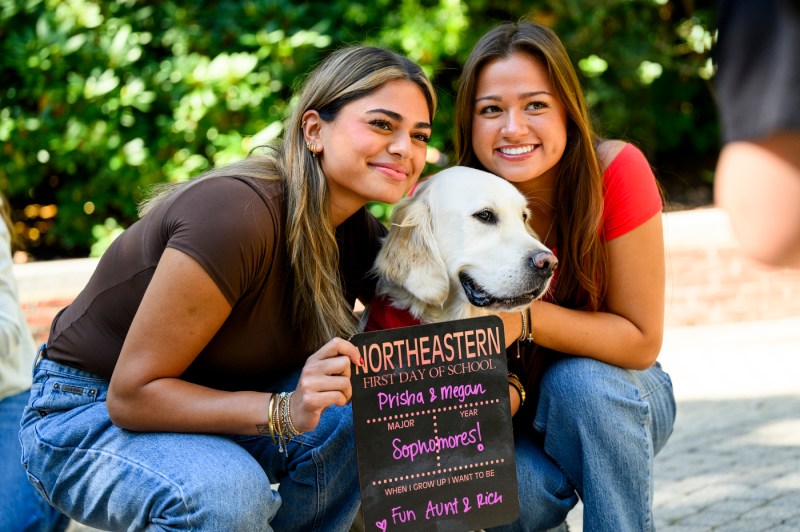 Students pose for a photo with a campus dog while holding up a "First Day of School" sign.