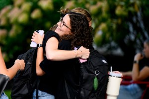 Two students embrace in a warm hug outdoors on campus. One of them has curly dark hair and glasses. Both are wearing dark clothes and backpacks and holding their phones in their hands.