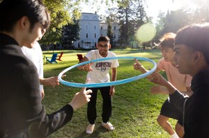 A group of college students participate in a team-building exercise on a sunny campus lawn, holding a large blue hoop together while standing in a circle.