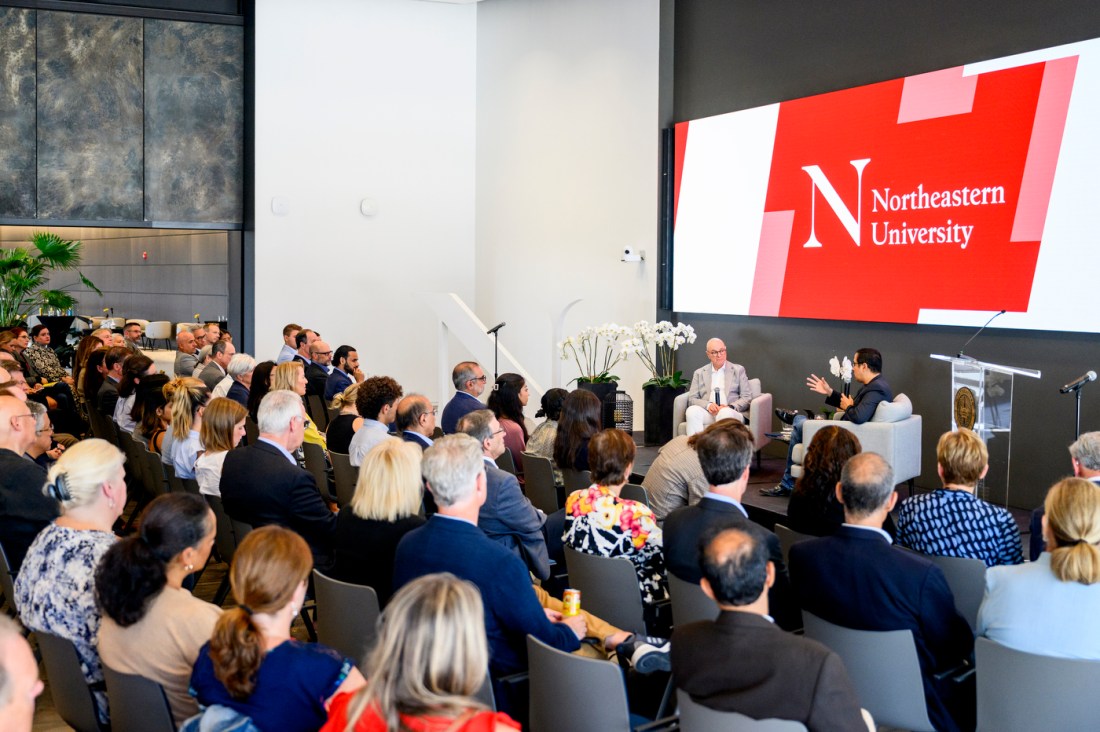 President Aoun and Spencer Fung sitting in grey arm chairs on a raised stage in front of rows of audience members. Behind them is a screen showing a red and white logo reading "Northeastern University". 