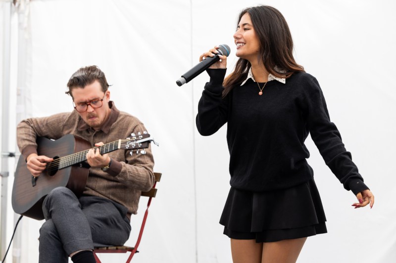Musical performers, including a female vocalist and a male guitarist, play music together in front of a white backdrop.