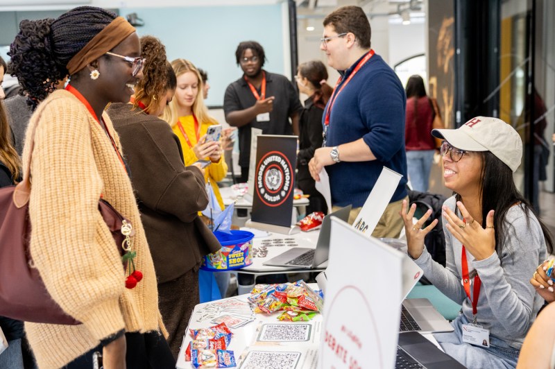 A black woman in a faded, light brown sweater talks to a light-skinned woman at a booth during Husky Fest.