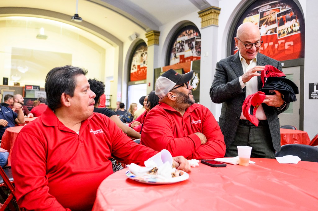 President Joseph Aoun, wearing a dark suit and glasses, stands and hands out red 'Northeastern' branded hats to facilities staff members wearing red shirts seated around a round table with a red tablecloth.