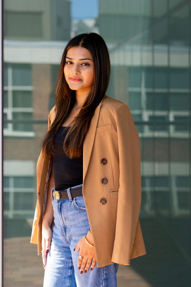 Khushi Shah, a dark-haired woman, shown posing for the camera in blue jeans and a brown blazer against a sleek backdrop.