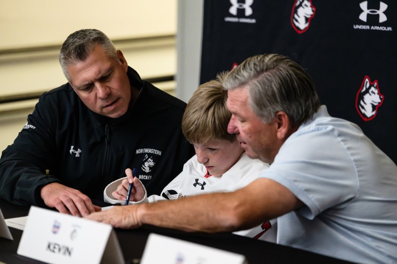 Eleven-year-old Mack Welby, who is sitting in between two adults and in front of a Northeastern banner, signs on to the Northeastern hockey team.