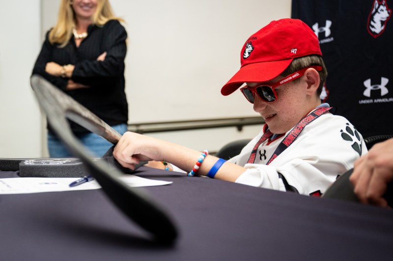 Mack Welby, who is wearing a Northeastern hat and sunglasses, smiles while looking at a hockey stick.