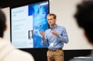 Baudouin Roos wearing a blue checkered shirt and orange glasses standing in front of audience members presenting. He is standing next to a projector screen and gesturing with his hands.