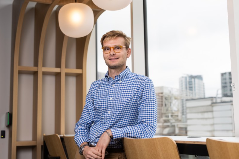 Baudouin Roos wearing a blue checkered shirt and orange glasses posing inside of a modern building with large windows.