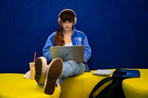 A student wearing headphones sits on a bench with her legs stretched out while working on a laptop.