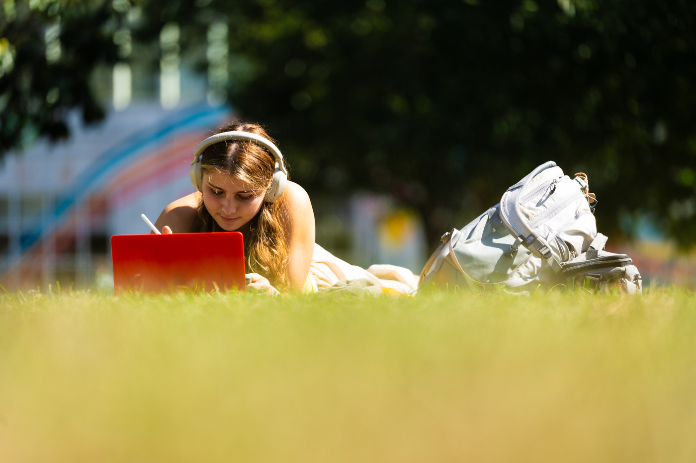 A student lays in the grass with their laptop open in front of them and their backpack next to them, studying at Centennial Common.