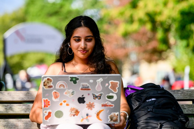 A student sits on a bench while working on a laptop, which is covered in stickers.